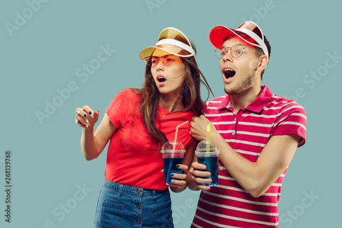 Beautiful young couple's half-length portrait isolated on blue studio background. Smiling woman and man in caps and sunglasses with drinks. Facial expression, summer, weekend concept. Trendy colors.