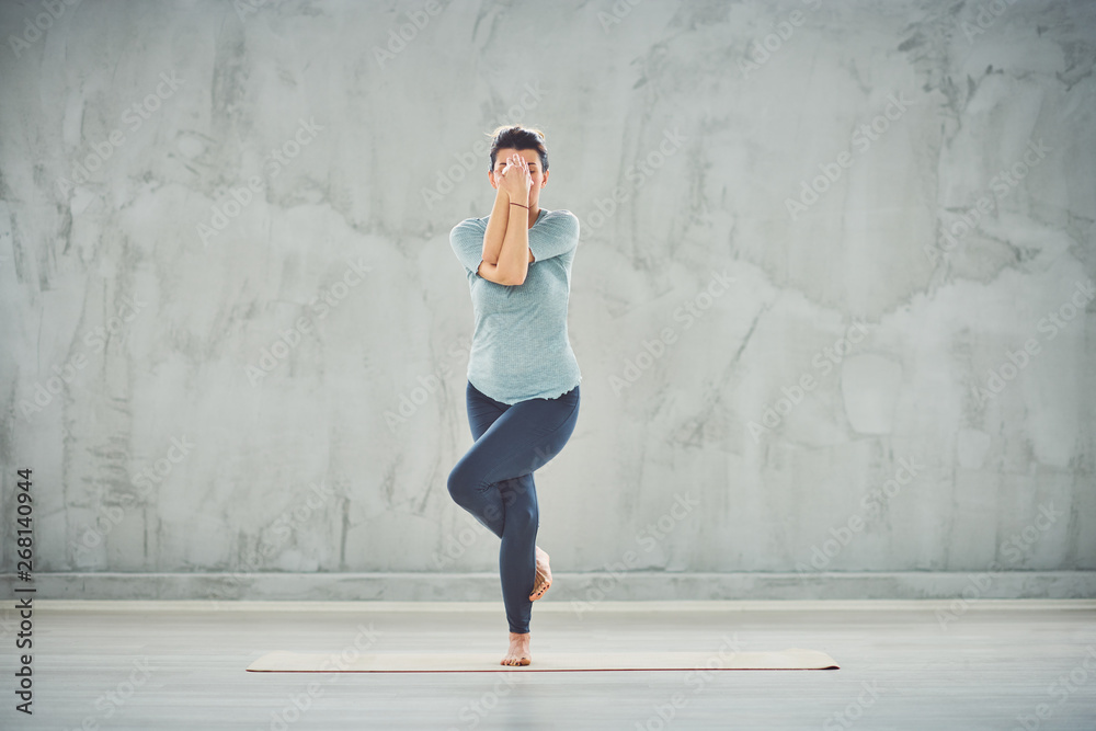 Full length of beautiful Caucasian brunette standing on the mat barefoot in Eagle yoga pose. Full length of beautiful Caucasian brunette standing on the mat barefoot in Eagle yoga pose.
