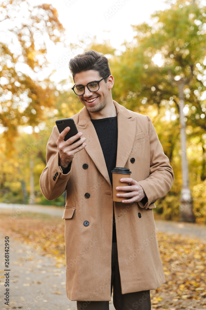 Image of happy young man wearing coat typing on smartphone and smiling in autumn park Image of happy young man wearing coat typing on smartphone and smiling in autumn park