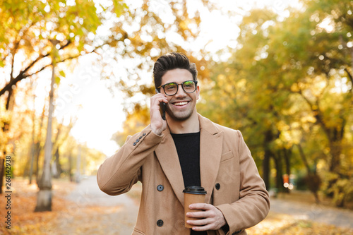 Image of handsome young man wearing coat talking on smartphone and smiling in autumn park