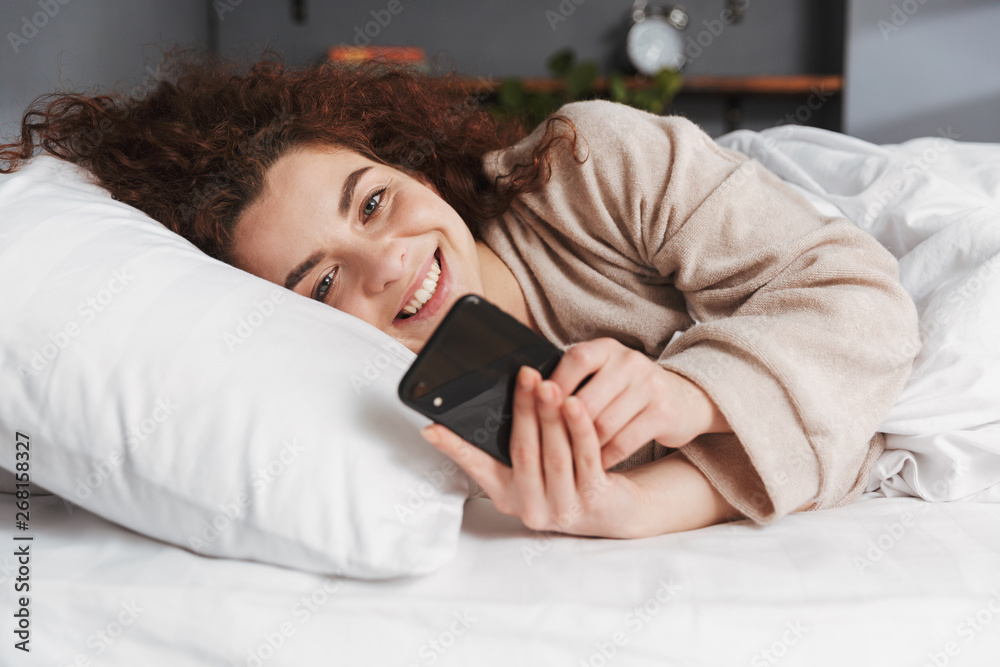 Photo of smiling young woman using smartphone while lying in bed on white linen at home Photo of smiling young woman using smartphone while lying in bed on white linen at home