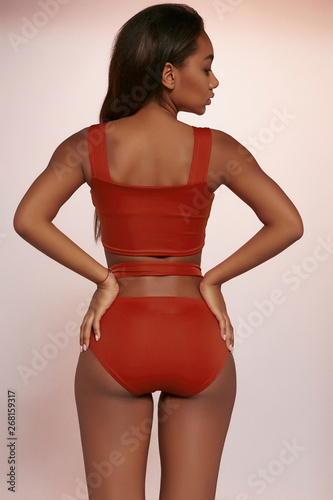 Three quarter back view shot of African lady, wearing terracotta red two-piece swimsuit with tank top and high-waisted panties. The girl is touching her tushie, looking to side on peachy background. photo