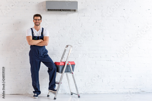 smiling repairman standing under air conditioner near stepladder and toolbox and looking at camera