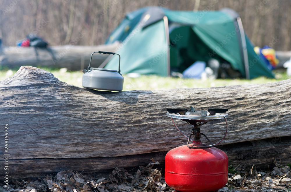 Camping gas bottle and metal kettle the camp background in the forest on sunny spring day. Tourist equipment for making tea and coffee on a gas during the journey. Focus on the big red gas bottle.