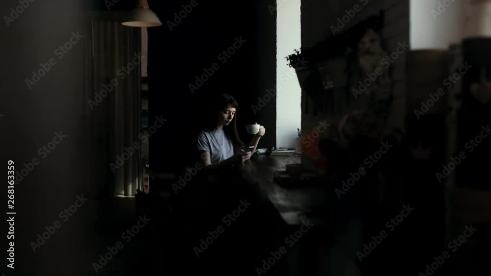 Girl sits by window, drinks coffee from a cup and typing message on phone in slow motion. Camera moves horizontally. Someone is spying on young woman who drinks sitting at table in cafe and texting Girl sits by window, drinks coffee from a cup and typing message on phone in slow motion. Camera moves horizontally. Someone is spying on young woman who drinks sitting at table in cafe and texting