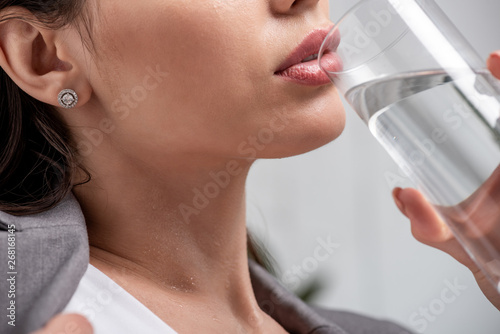 cropped view of young woman suffering from heat and drinking water from glass on grey