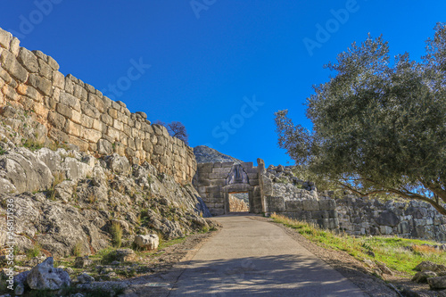 View of Lions Gate at ancient Mycenae Greece from down the hill showing the sidewalk - shaded by an olive tree - leading through the opening and the hill behind it