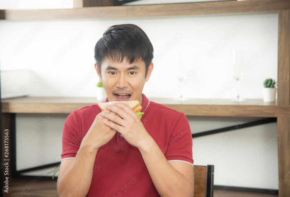 The business smiling man with casual red t-shirt having breakfast and eating sandwich, Young man cooking food and drink in the loft style kitchen room The business smiling man with casual red t-shirt having breakfast and eating sandwich, Young man cooking food and drink in the loft style kitchen room