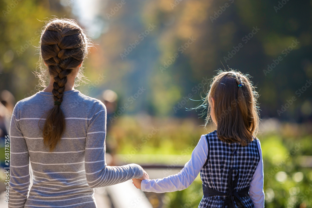 Fototapeta premium Back view of child girl and mother in dresses together holding hands on warm day outdoors on sunny background.