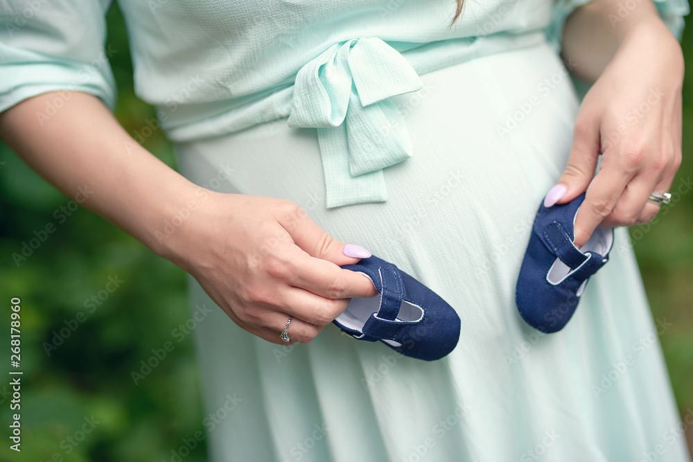 Close up of beautiful young Caucasian woman in her last trimester of pregnancy wearing a mint vaporous summer dress and holding a pair of blue baby shoes revealing the gender of the baby as a boy