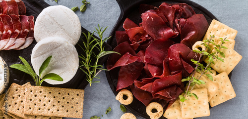 banner of Italian bresaola served sliced on a tray on a table with white wine, crackers, grissini and taralli with aromatic herbs on a blue linen festive tablecloth. photo