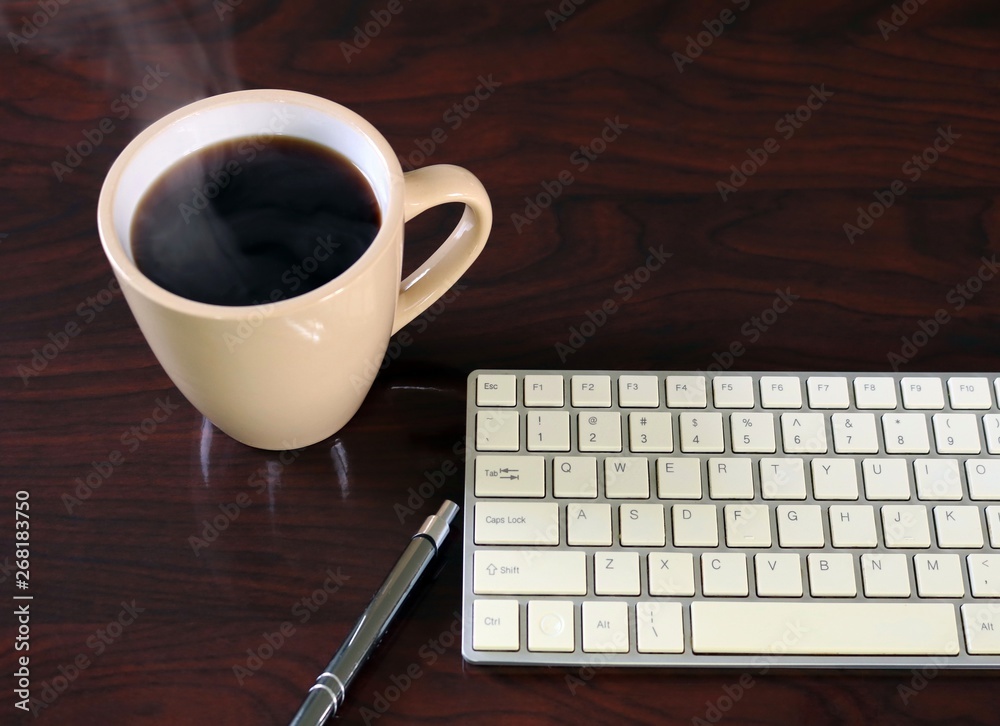 Steaming hot cup of coffee beside keyboard on dark wood grain desk Steaming hot cup of coffee beside keyboard on dark wood grain desk