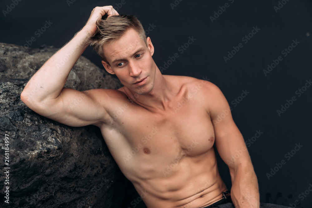 Portrait of young man with naked torso siting on rocky beach with black sand.