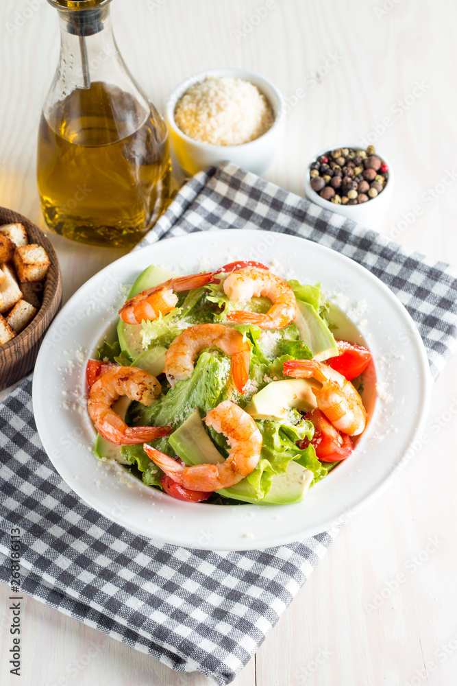 Fresh shrimp salad made of tomato, ruccola, avocado, prawns, chicken breast, arugula, crackers and spices. Caesar salad in a white, transparent bowl on wooden background