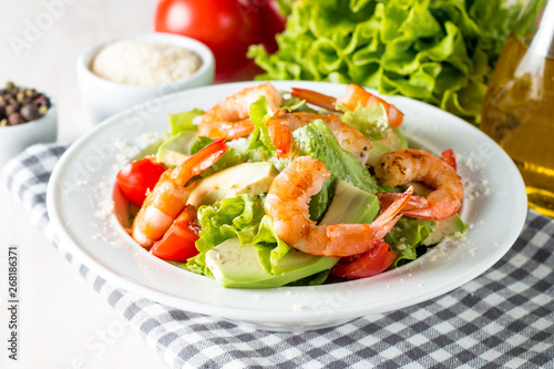 Fresh shrimp salad made of tomato, ruccola, avocado, prawns, chicken breast, arugula, crackers and spices. Caesar salad in a white, transparent bowl on wooden background