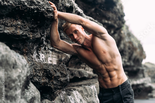 Sexy fitness male model in black pants and shirtless posing on black sandy beach.