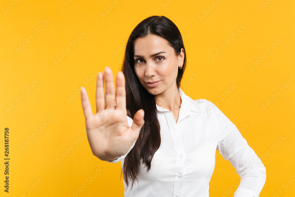 Portrait of beautiful young woman in white shirt looking camera showing stop gesture with palm isolated on bright yellow orange wall background in studio. People lifestyle concept. Mock up copy space. Portrait of beautiful young woman in white shirt looking camera showing stop gesture with palm isolated on bright yellow orange wall background in studio. People lifestyle concept. Mock up copy space.