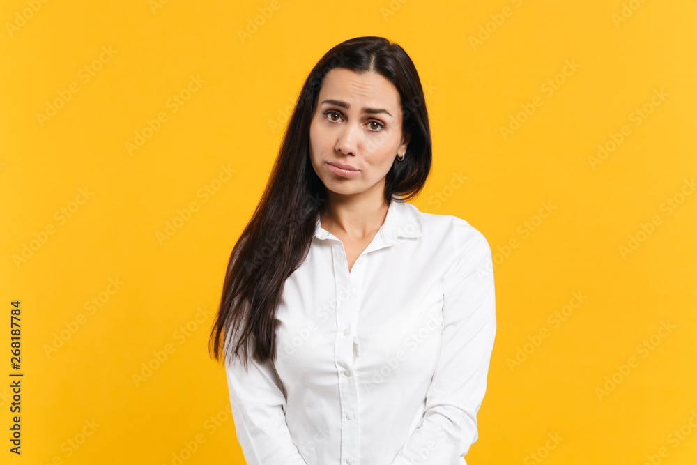 Fototapeta premium Portrait of upset offended young woman in white casual shirt standing and looking camera isolated on bright yellow orange wall background in studio. People lifestyle concept. Mock up copy space.