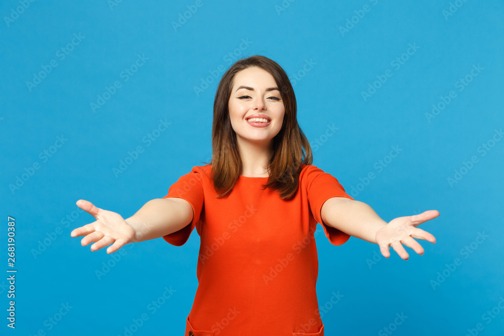 Beautiful young woman wearing red orange dress outstretched hand to camera standing posing isolated over blue wall background, studio portrait. People lifestyle fashion concept. Mock up copy space. Beautiful young woman wearing red orange dress outstretched hand to camera standing posing isolated over blue wall background, studio portrait. People lifestyle fashion concept. Mock up copy space.