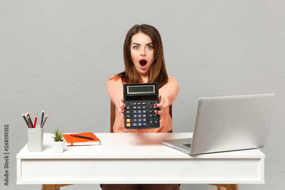 Young shocked woman in pastel clothes keeping mouth open, hold calculator, work at desk with pc laptop isolated on gray background. Achievement business career lifestyle concept. Mock up copy space. Young shocked woman in pastel clothes keeping mouth open, hold calculator, work at desk with pc laptop isolated on gray background. Achievement business career lifestyle concept. Mock up copy space.