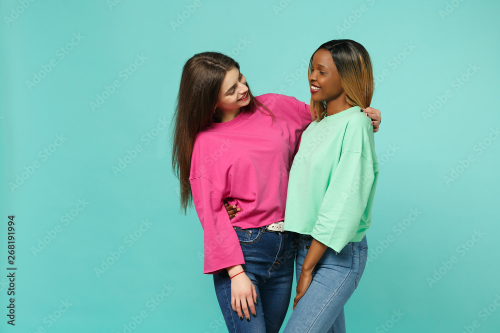 Two young women friends european and african american in pink green clothes standing posing isolated on blue turquoise wall background, studio portrait. People lifestyle concept. Mock up copy space. Two young women friends european and african american in pink green clothes standing posing isolated on blue turquoise wall background, studio portrait. People lifestyle concept. Mock up copy space.