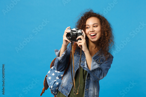 Young african american girl teen student in denim clothes backpack hold camera isolated on blue wall background studio portrait. Education in high school university college concept. Mock up copy space photo