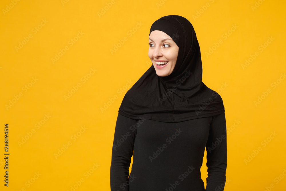 Fototapeta premium Smiling beautiful young arabian muslim woman in hijab black clothes looking aside camera isolated on yellow wall background, studio portrait. People religious lifestyle concept. Mock up copy space.