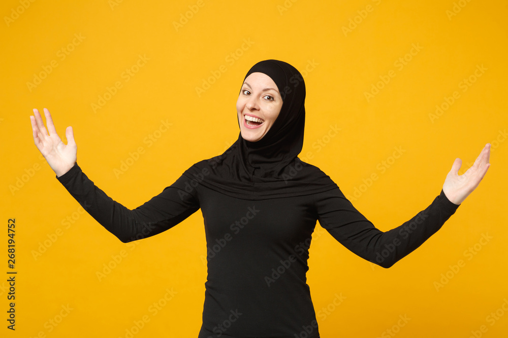 Smiling young arabian muslim woman in hijab black clothes showing pointing copy space with hands finger isolated on yellow wall background, studio portrait. People religious lifestyle concept. Mock up Smiling young arabian muslim woman in hijab black clothes showing pointing copy space with hands finger isolated on yellow wall background, studio portrait. People religious lifestyle concept. Mock up