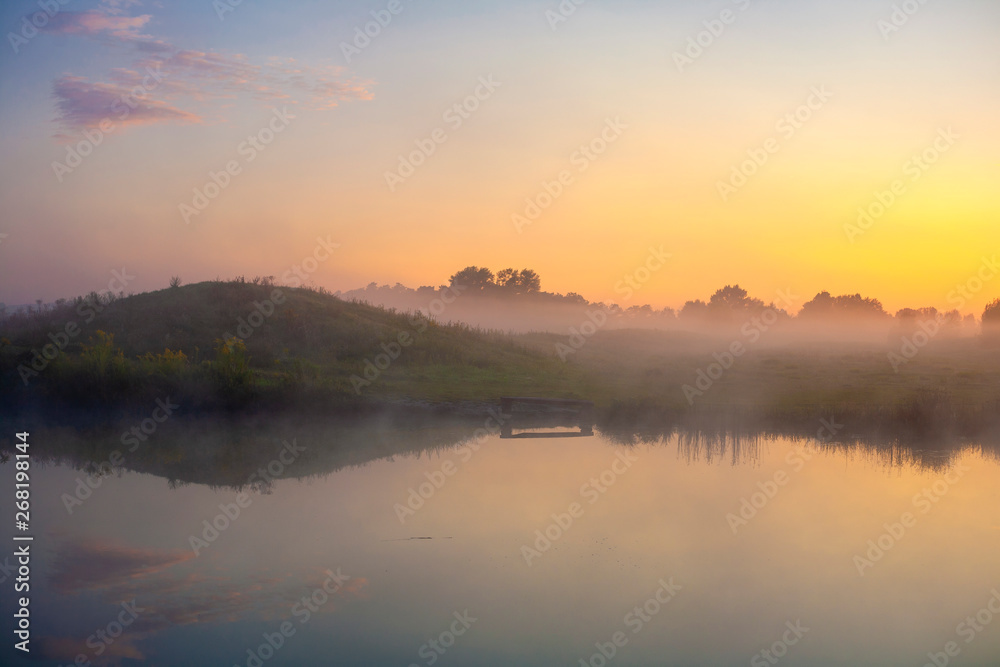 Picturesque misty sunrise over a lake with a bench and a park