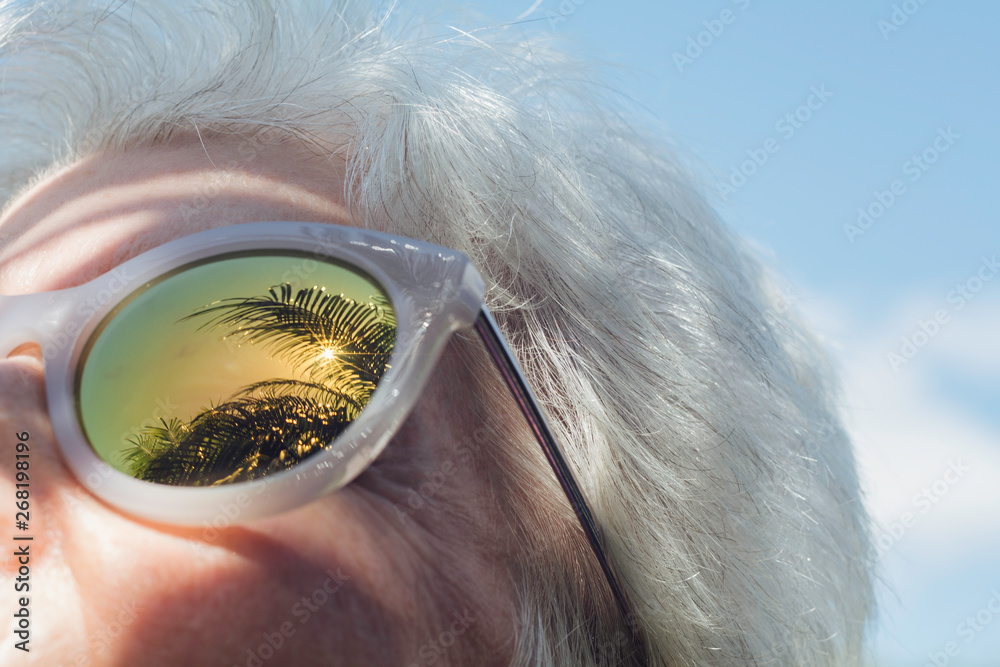 © Kristin Duvall/Stocksy - Senior woman with palm trees reflected in sunglasses
