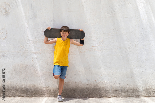 Fototapeta Naklejka Na Ścianę i Meble -  Young smiling boy leaning on yellow wall holding a skateboard while looking camera in a bright day
