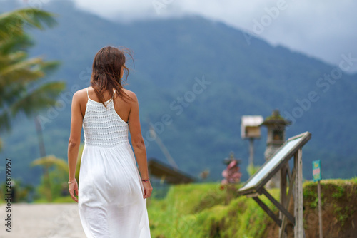 A tanned girl in a white dress walks forward on the road and look on a sign. The view from the back. In the background, a mountain in the fog. Close up