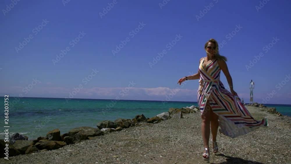 charming blonde woman is running over seashore in sunny day, playing with her long dress