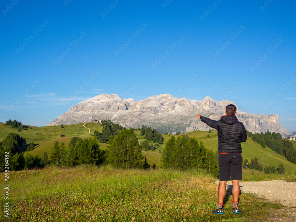 A person admires the fantastic Dolomites in Italy during the summer period. The massive Sella in the background. Hiker photographed from behind. Dolomites a Unesco World Heritage A person admires the fantastic Dolomites in Italy during the summer period. The massive Sella in the background. Hiker photographed from behind. Dolomites a Unesco World Heritage