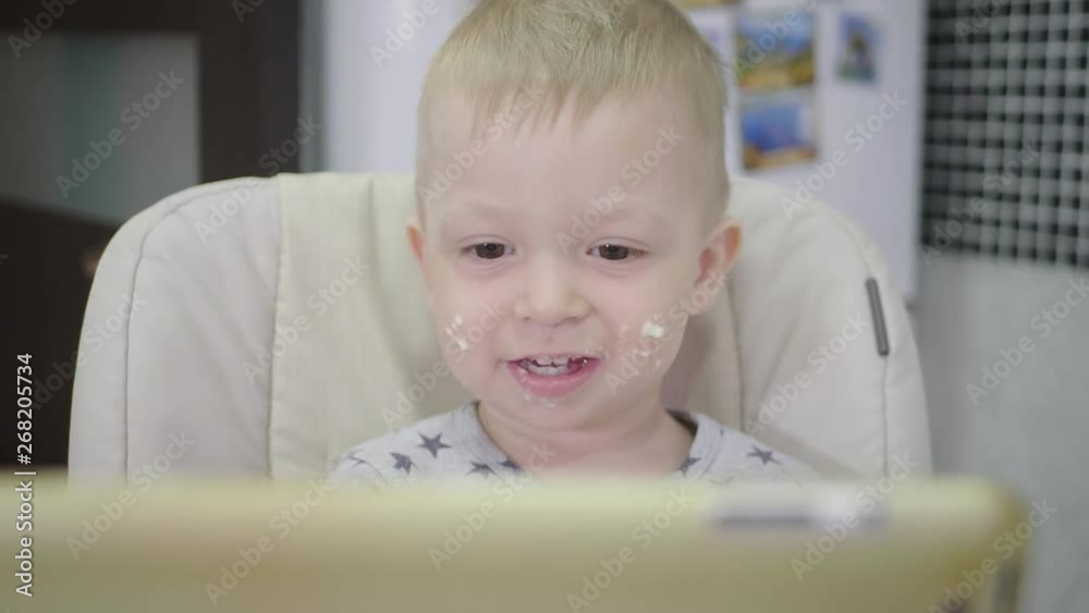 Cute little boy, eating porridge for breakfast at home, while watching cartoon on tablet. Children and technology or healthy food for kid concept. Cute little boy, eating porridge for breakfast at home, while watching cartoon on tablet. Children and technology or healthy food for kid concept.