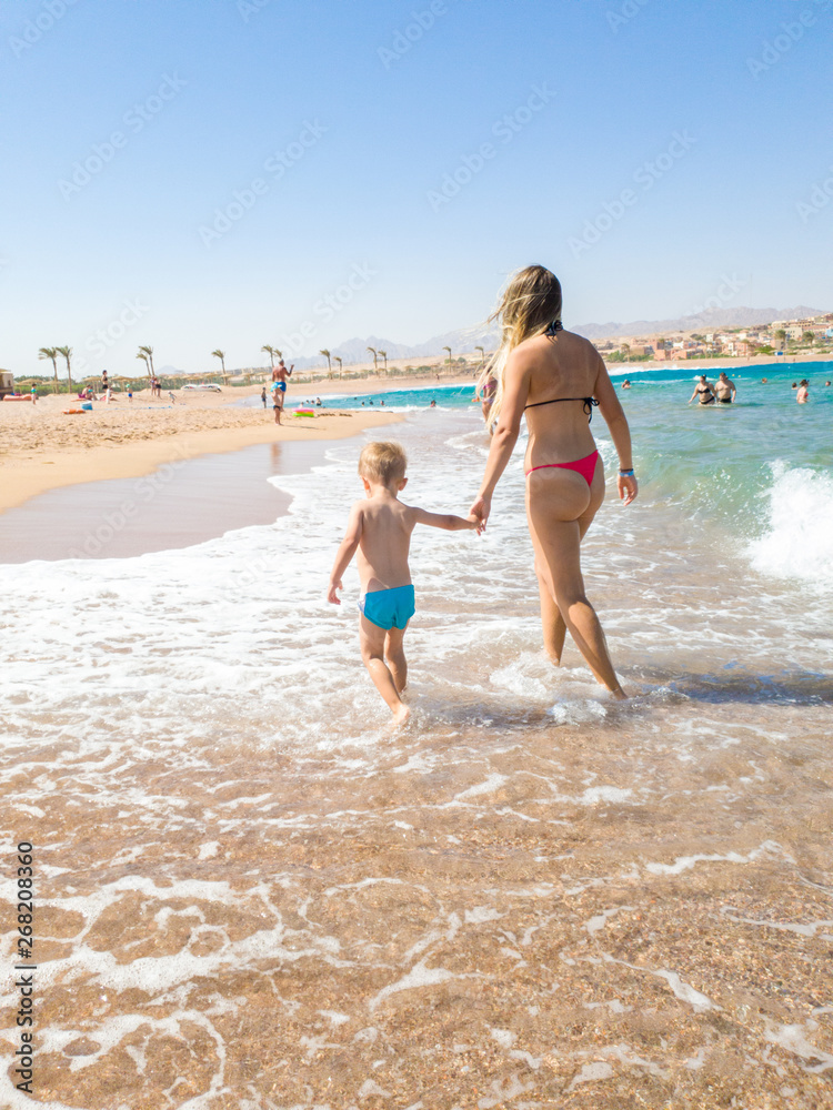 3 years old little boy holding his young mother by hand and walking on the sea beach. Family relaxing and having good time during summer holiday vacation.