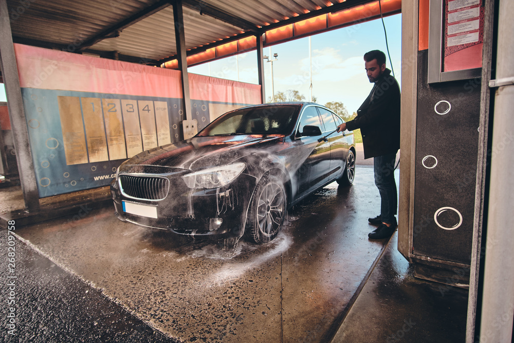 Smart trendy man in jeans and blaser is washing his own car at car washing station.