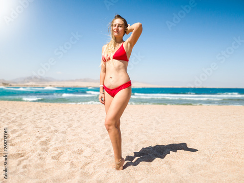 Beautiful slim young woman with long hair wearing sexy red bikini posing on the beach against sea waves and blue sky