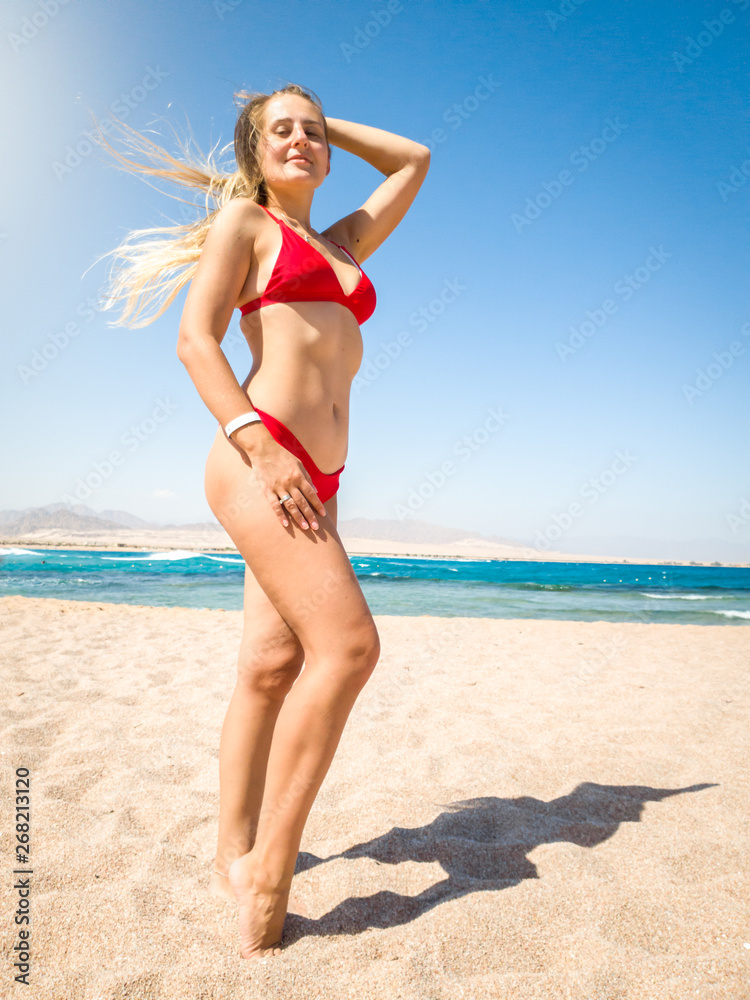 Beautiful slim young woman with long hair wearing sexy red bikini posing on the beach against sea waves and blue sky