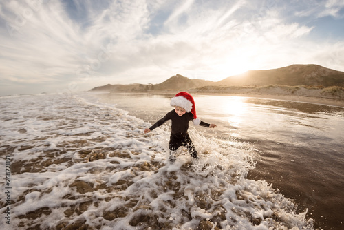Toddler boy wearing red hat splashing in waves at beach at dusk