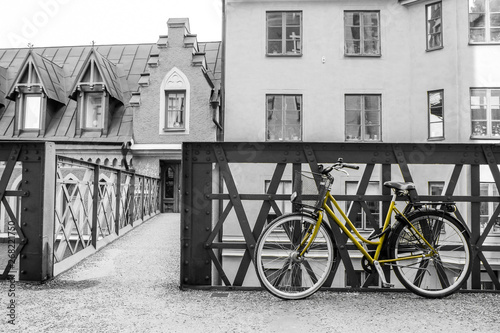 A picture of a lonely yellow bike standing in the typical street in Stockholm by the bridge to a house. The bike looks to be modern in a retro style. The background is black and white. 