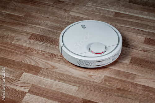 white robotic vacuum cleaner runs on laminate floor. Robot controlled by voice commands for direct cleaning. Modern smart appliance for cleaning house