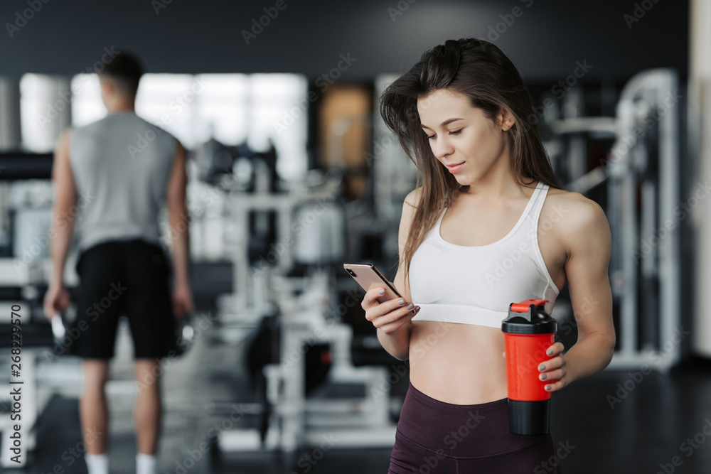 Beautiful young sportswoman in sportswear and earphones is holding a bottle of water, listening to music using a phone and smiling, in the gym.