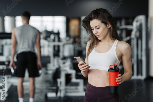 Fototapeta Naklejka Na Ścianę i Meble -  Beautiful young sportswoman in sportswear and earphones is holding a bottle of water, listening to music using a phone and smiling, in the gym.