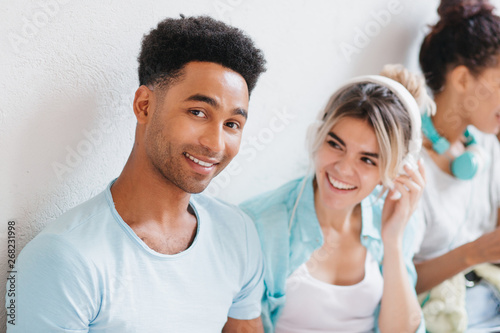 Cheerful young man with brown skin and big smile posing beside white wall. Portrait of glad black-haired guy and laughing girl in big earphones enjoying music.