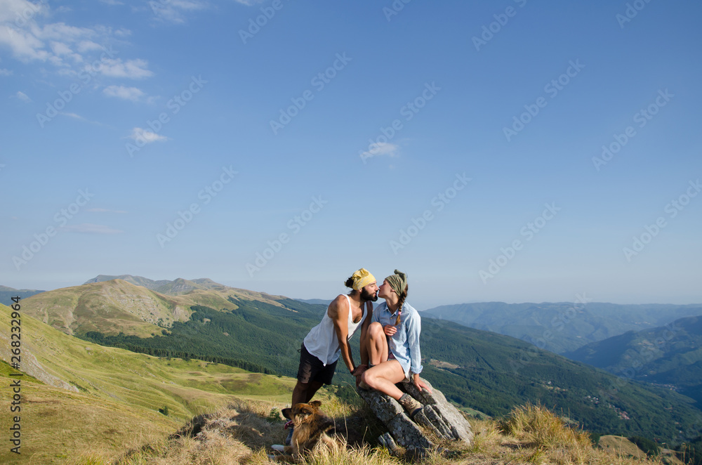 Obraz premium a couple is kissin on top of a mountains with their dog sit at their feet, and the landscape in background