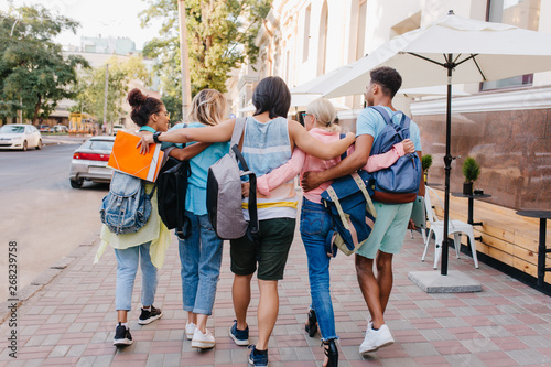 Portrait from back of students with stylish backpacks walking down the street after lectures in university. Tall brunette young man embracing girls while spending time with them outdoot.