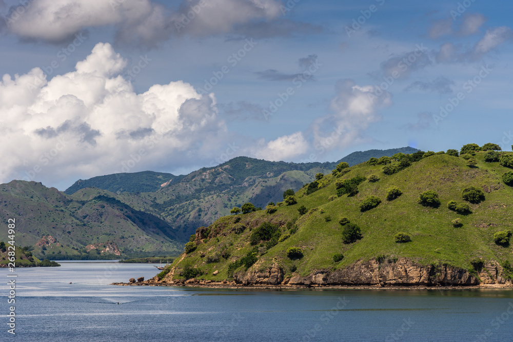 Komodo Island, Indonesia - February 24, 2019: Green mountains descending into sea water under blue sky with cloudscape, part of Komodo National Park.