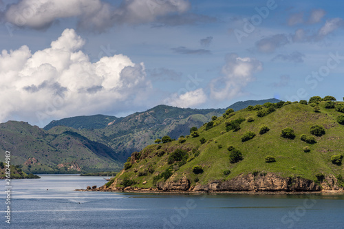 Komodo Island, Indonesia - February 24, 2019: Green mountains descending into sea water under blue sky with cloudscape, part of Komodo National Park. © Klodien