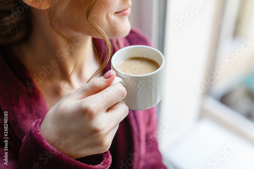 Morning coffee. Woman holding white Cup of black coffee sitting by the window. Young girl enjoys hot invigorating coffee in the morning